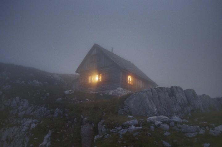 A wooden lodge with illuminated windows sits on a rocky, grassy hillside shrouded in fog.