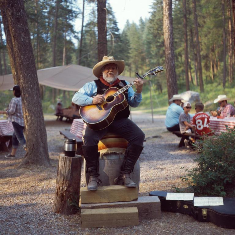 A man with a white beard and a cowboy hat plays an acoustic guitar while sitting on a barrel in a forest.