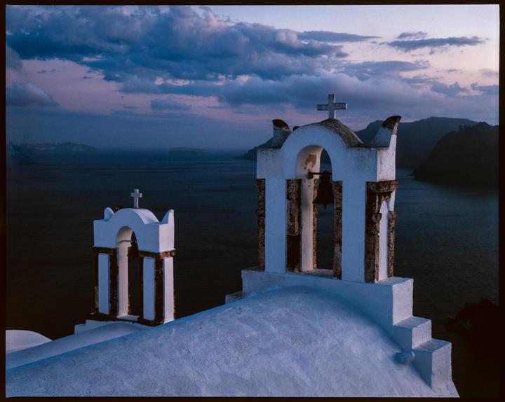Two white bell towers with crosses stand on a rooftop overlooking a body of water and mountains under a cloudy sky at dusk.