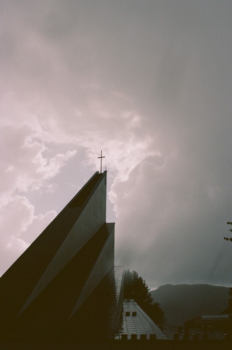 A modern church with a cross on its spire stands against a cloudy sky, with mountains in the background.