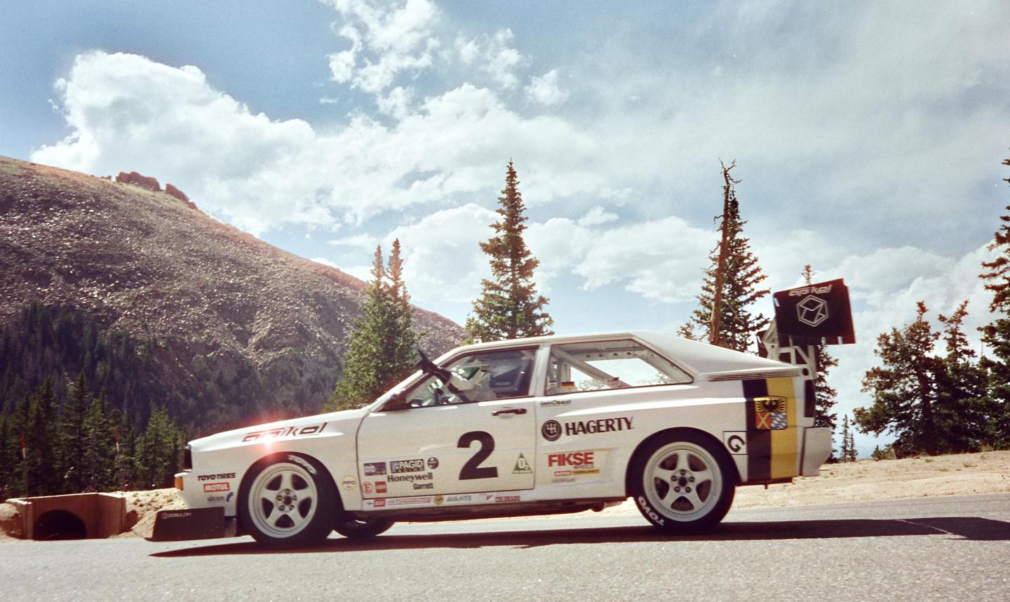 A white race car with the number two on its side is parked on a road with a mountain and pine trees in the background.