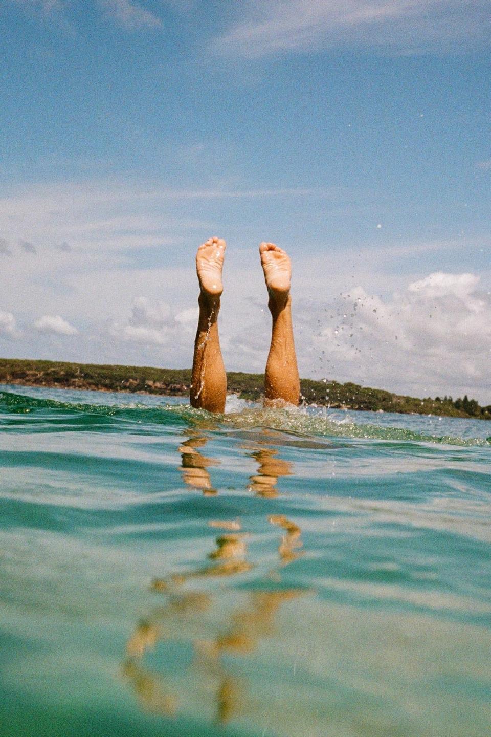 A person's legs and feet are visible above the water as they dive into a body of water under a blue sky.