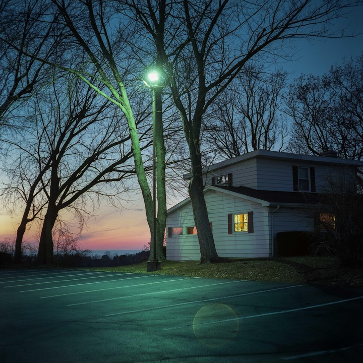 A house with illuminated windows stands next to a parking lot and bare trees under a colorful sky at dusk.