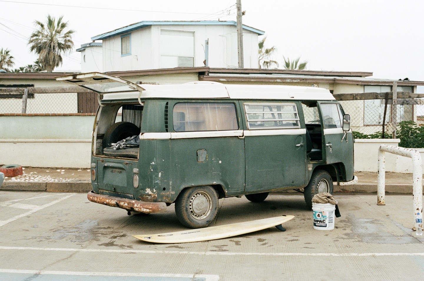 A green van with its back hatch open is parked on a paved surface next to a surfboard and a white bucket.