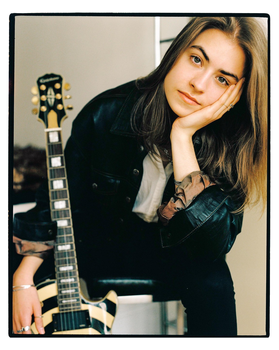 A young woman with long hair and a leather jacket poses with a striped electric guitar.