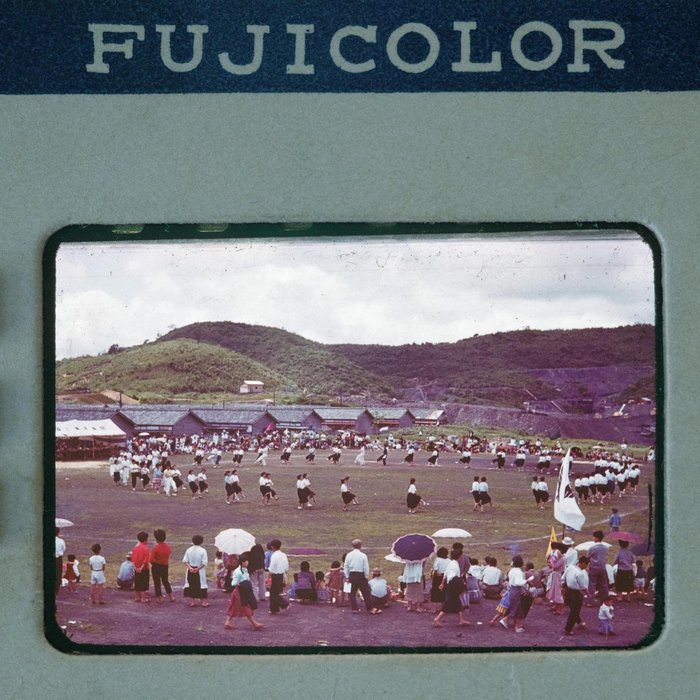 A large group of people are gathered on a grassy field with hills and buildings in the background.