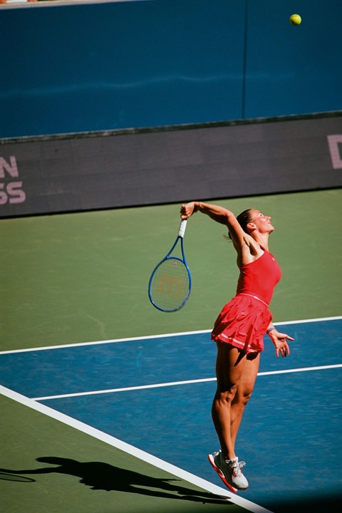 A woman in a red tennis outfit is captured mid-air during a serve on a green and blue tennis court.