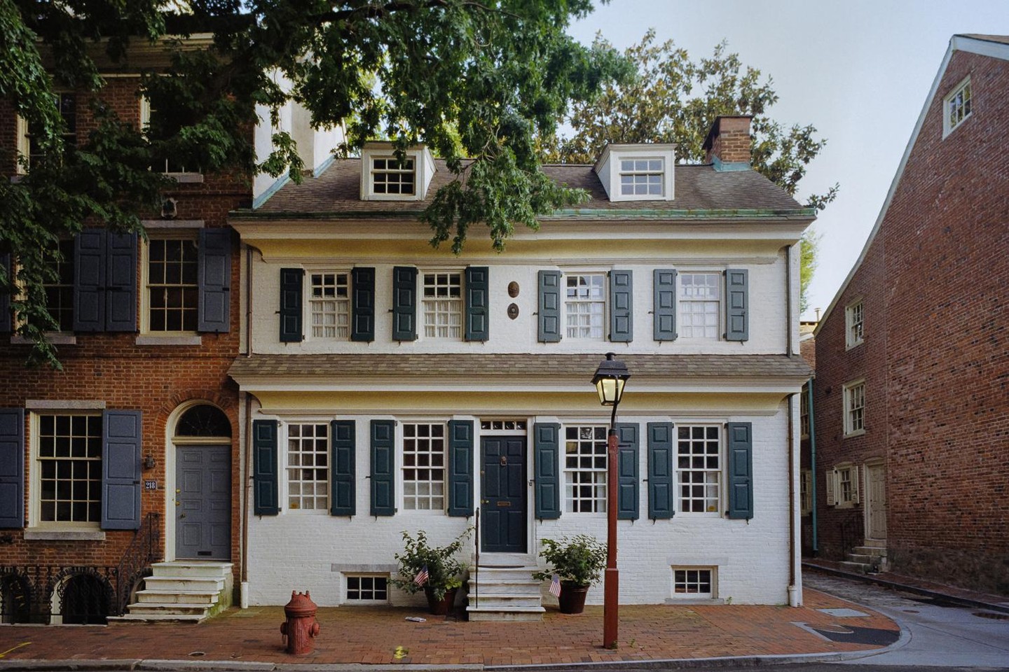 A white house with dark green shutters stands next to a brick house with blue shutters on a street.