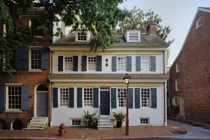 A white house with dark green shutters stands next to a brick house with blue shutters on a street.