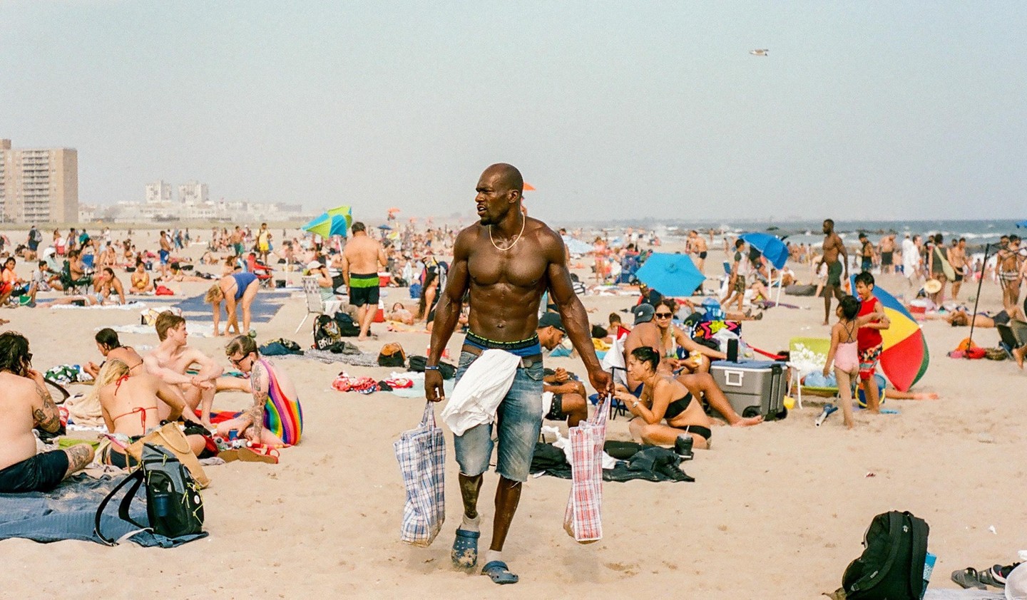 A muscular man walks across a crowded beach carrying two bags, with many people relaxing on the sand and buildings in the background.