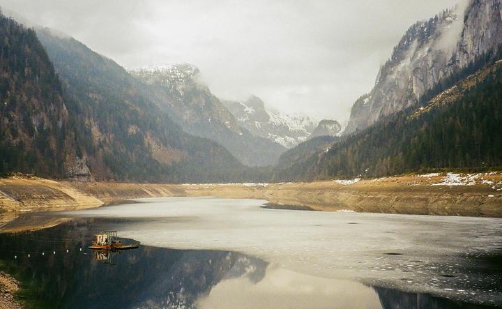 A partially frozen lake with a small boat is surrounded by mountains with some snow and evergreen trees under a cloudy sky.
