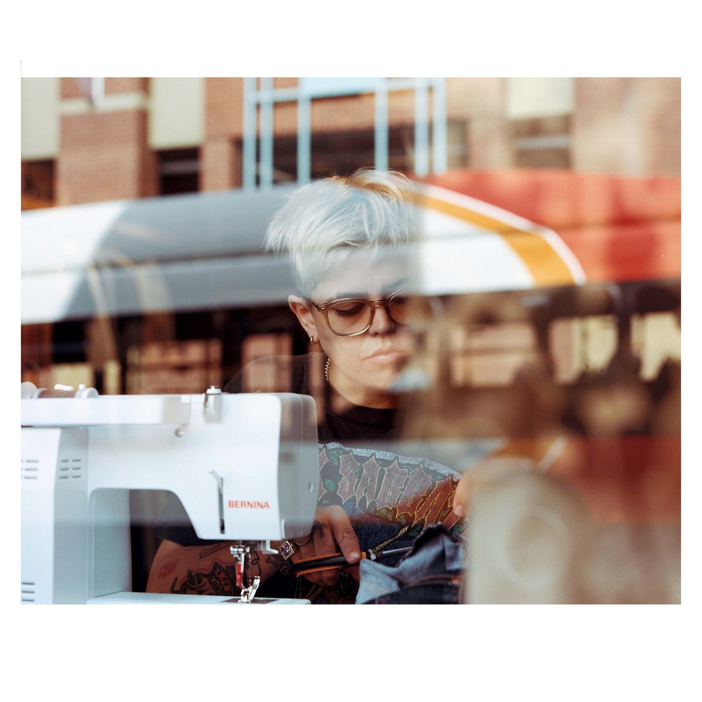 A woman with short blonde hair and glasses is seen through a window, sewing fabric on a sewing machine.