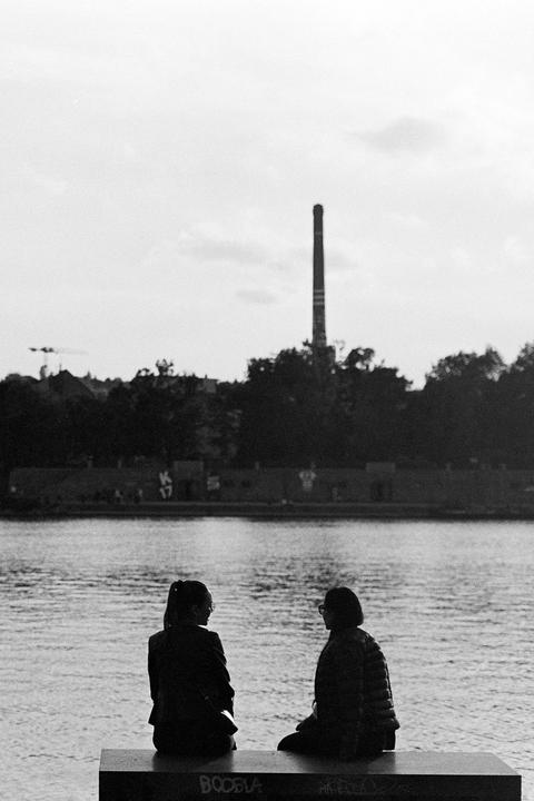 Two women sit on a bench by a river with a tall chimney in the background.