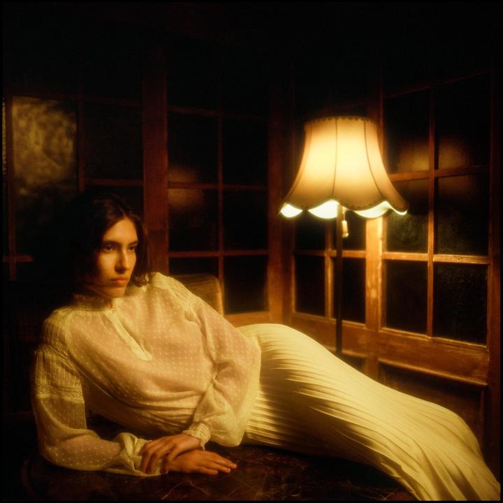A woman in a sheer blouse and pleated skirt is reclining next to a floor lamp in a dimly lit room.