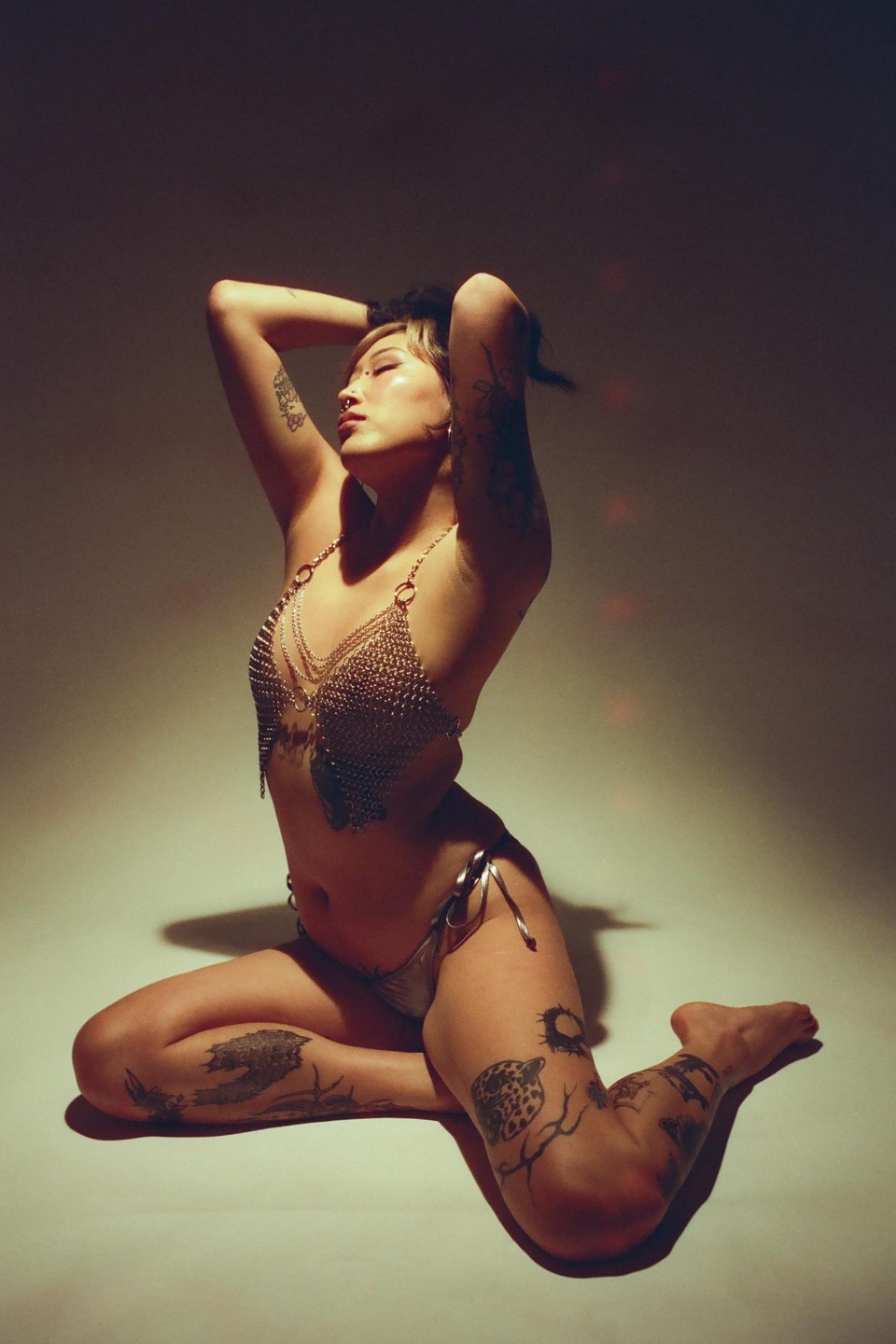 A woman with tattoos on her arms and legs kneels on the floor, wearing a chainmail top and bikini bottom, with her arms raised behind her head.