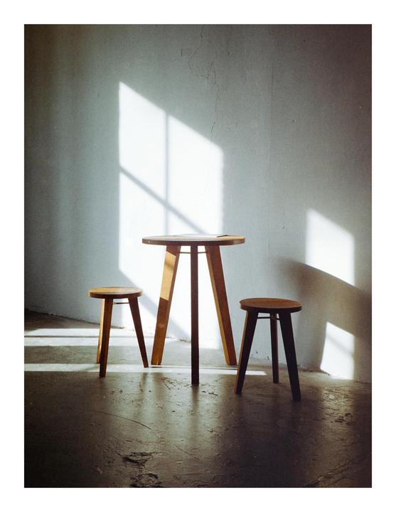 Sunlight casts shadows on a wall and floor behind a wooden table and two stools.