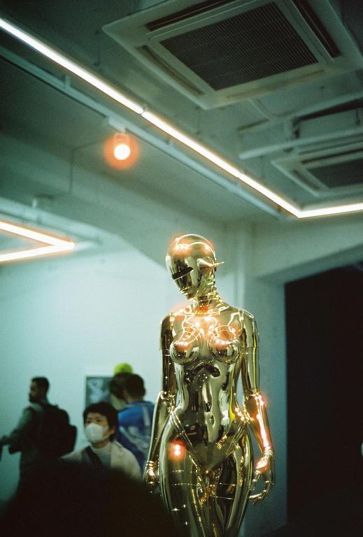 A golden robot sculpture stands in a room with people in the background.