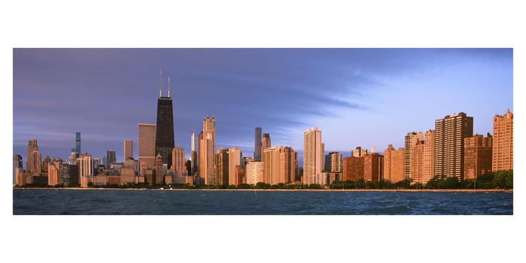 A panoramic view of the Chicago skyline at sunrise, with buildings illuminated by golden light and a body of water in the foreground.