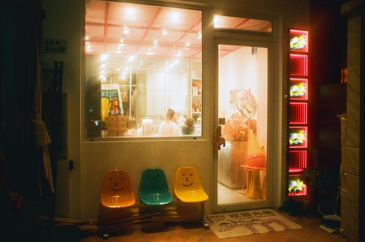 Three colorful chairs with faces are lined up outside a brightly lit storefront at night.