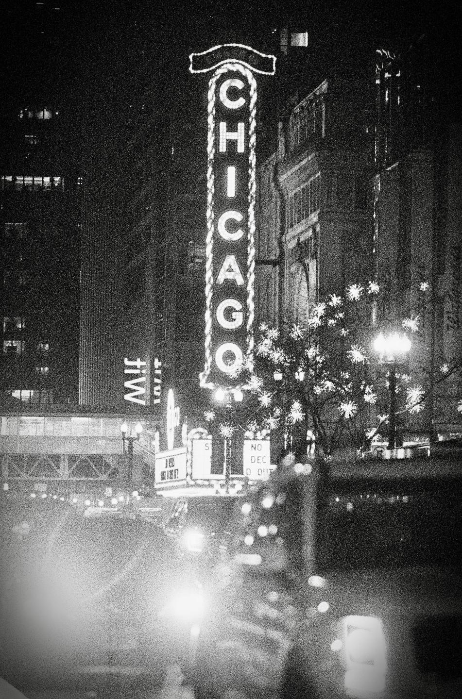 A tall neon sign spelling "CHICAGO" stands prominently at night, with blurred lights and buildings in the background.