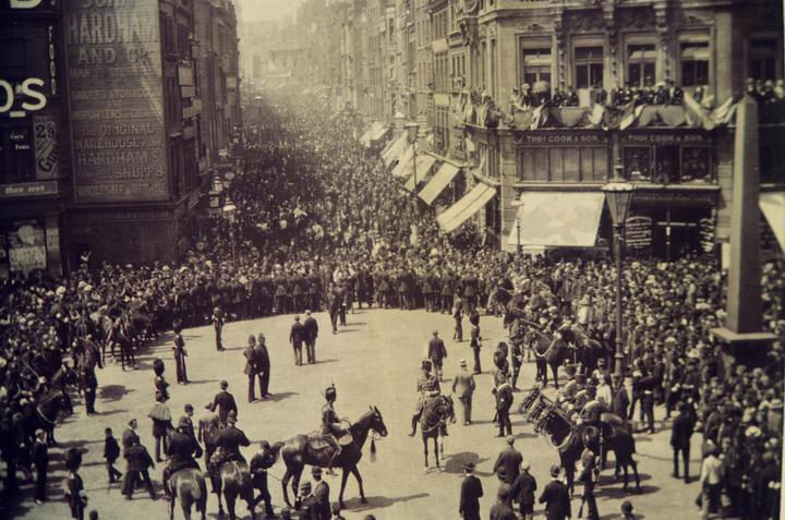 A large crowd of people and soldiers on horseback fill a city street lined with buildings.
