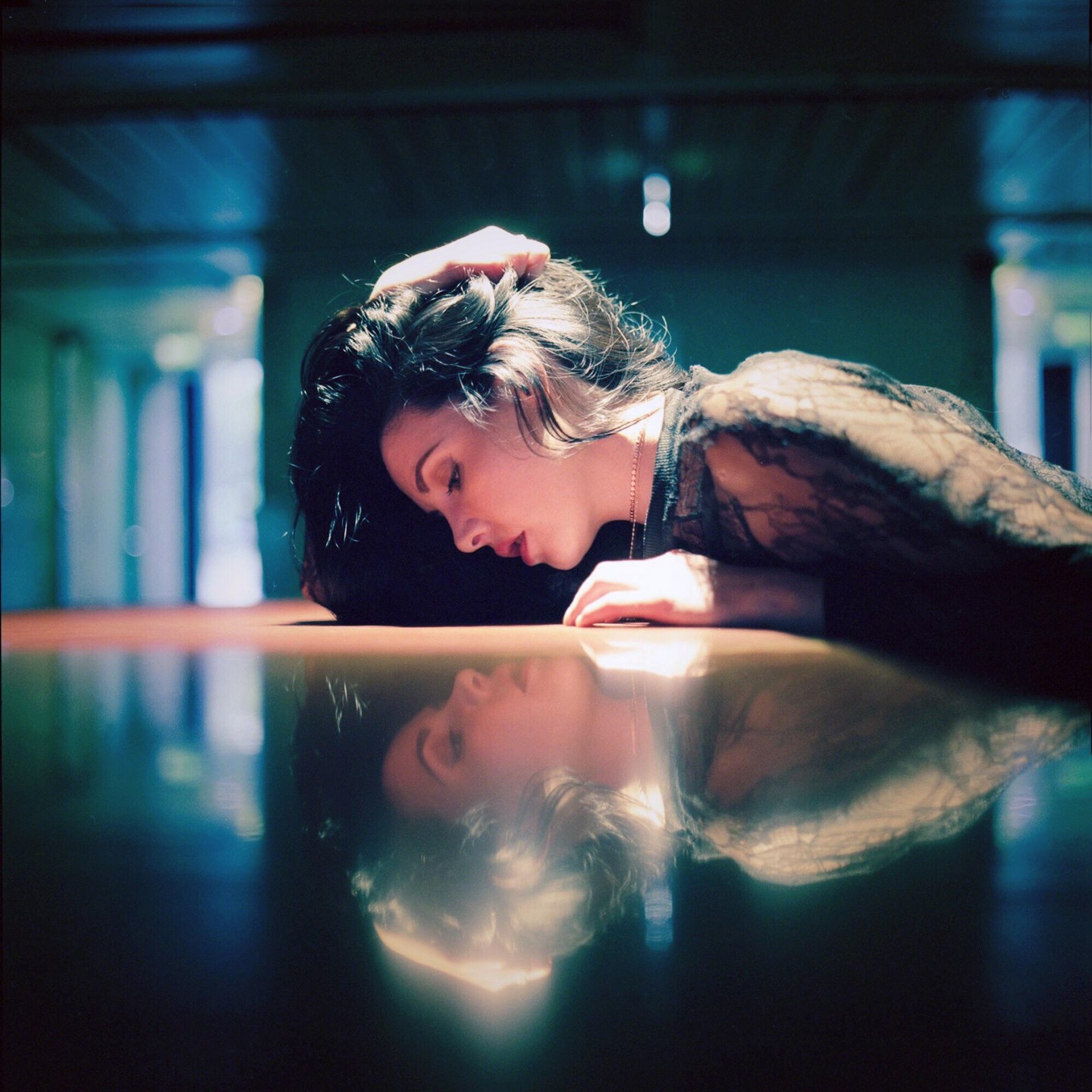 A woman with dark hair rests her head on a reflective surface, with her reflection visible below.