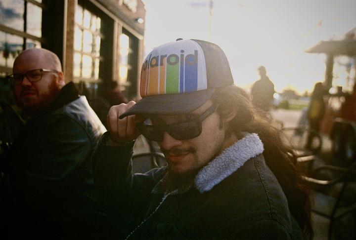 A man wearing a hat and sunglasses is looking down while another man with glasses is visible in the background.