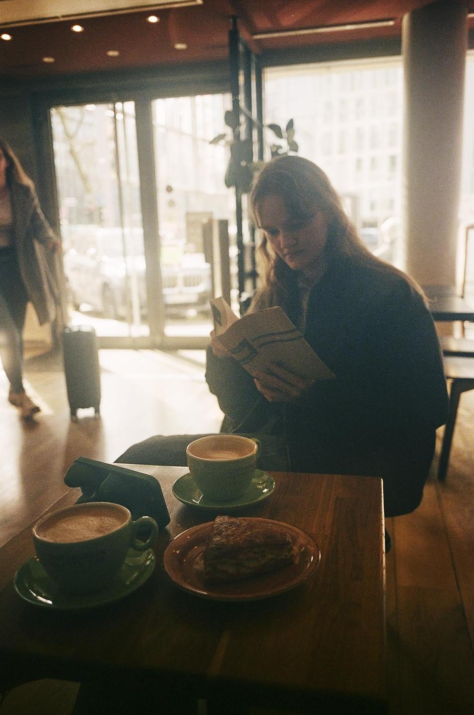 A woman reads a book at a cafe table with two cups of coffee and a pastry.