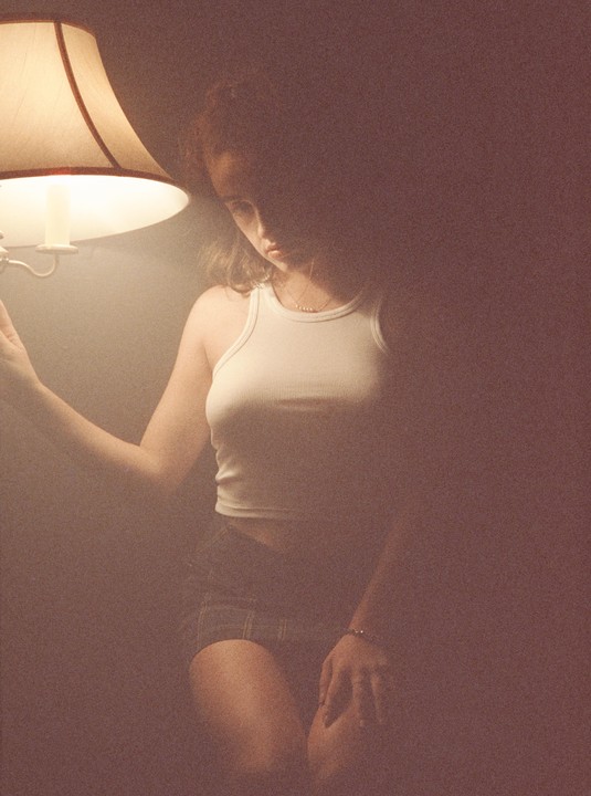 A woman in a white tank top and plaid shorts is illuminated by a lamp.