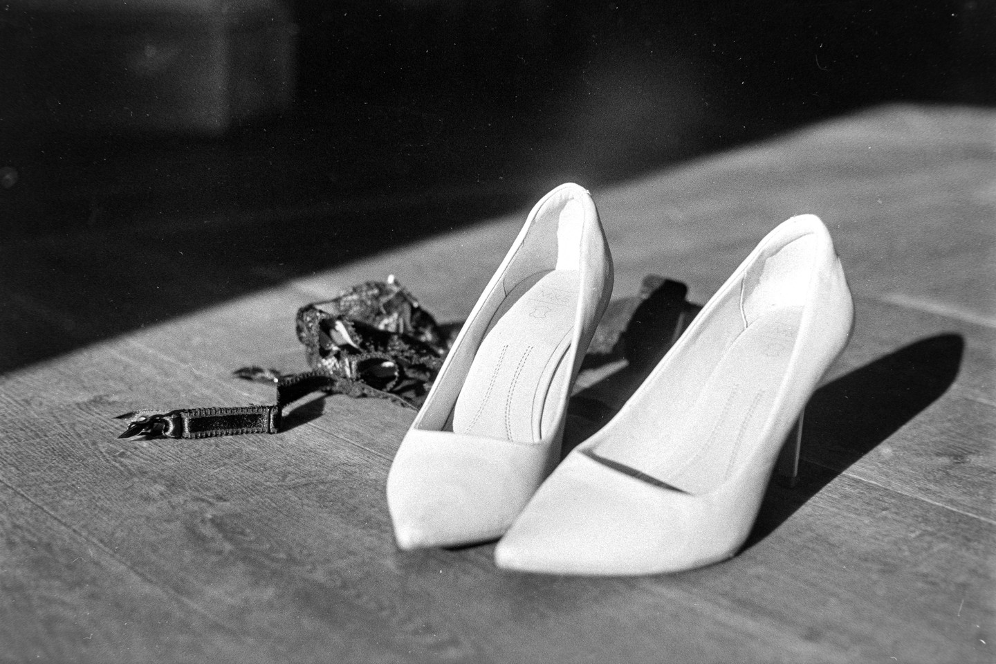 Two high-heeled shoes and a garter belt are on a wooden floor with a strong shadow.