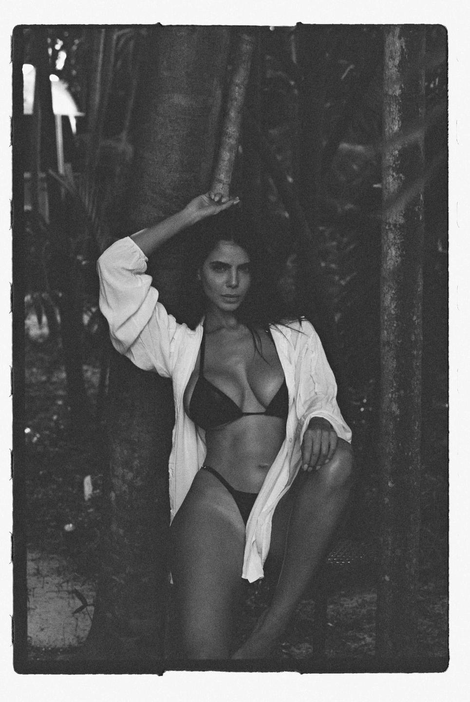A woman in a bikini and open shirt poses next to a tree.