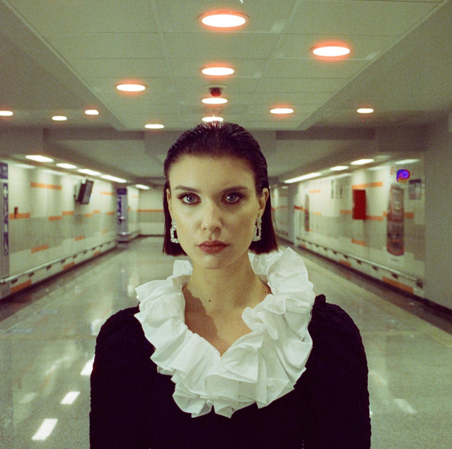 A woman with short hair and a ruffled collar stands in a brightly lit underground tunnel.