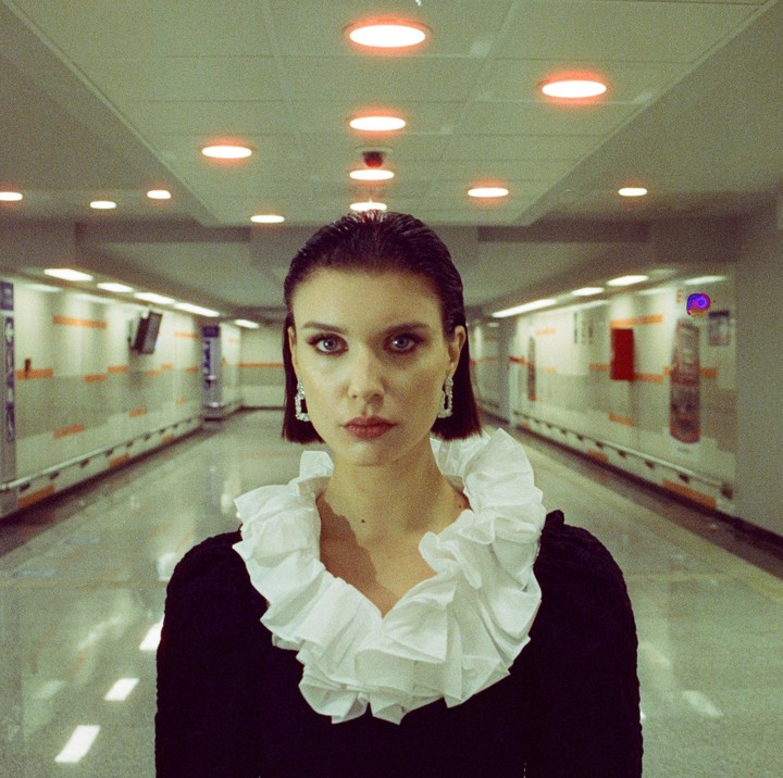 A woman with short hair and a ruffled collar stands in a brightly lit underground tunnel.