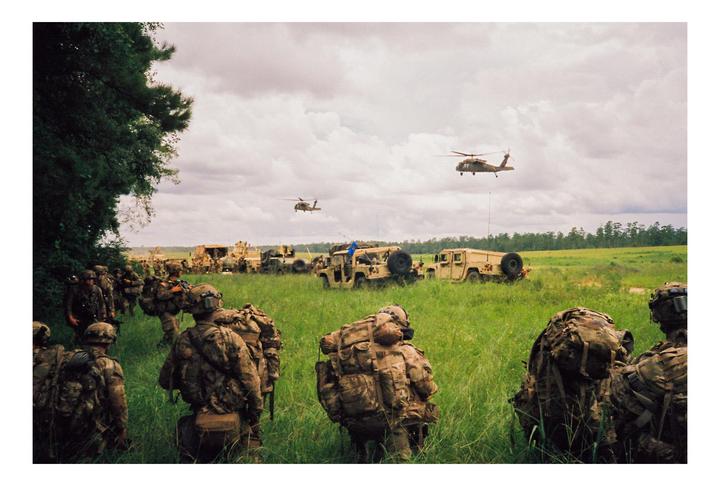 Soldiers with large backpacks stand in a grassy field with military vehicles and helicopters under a cloudy sky.