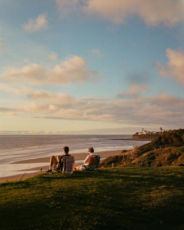 Two men sit on a grassy cliff overlooking a beach and ocean at sunset.