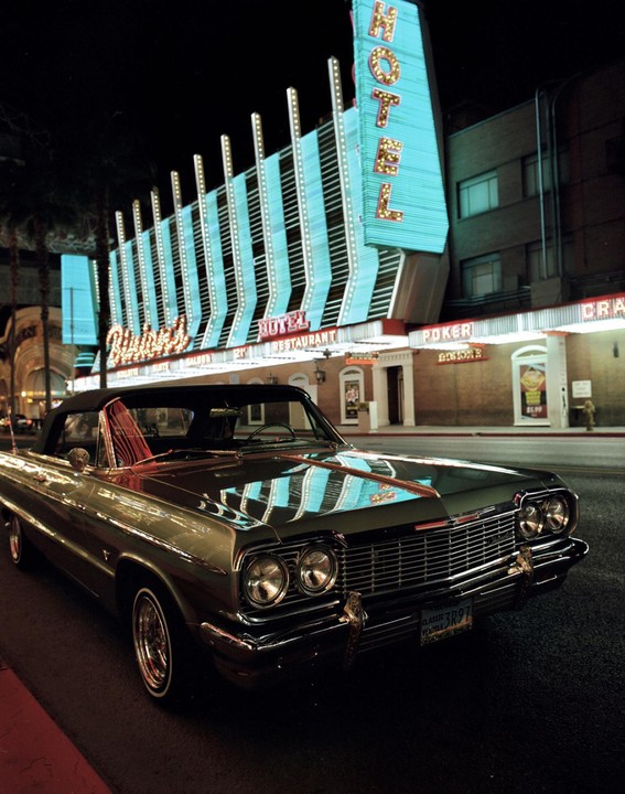 A classic car is parked on a street at night in front of a hotel with a large neon sign.