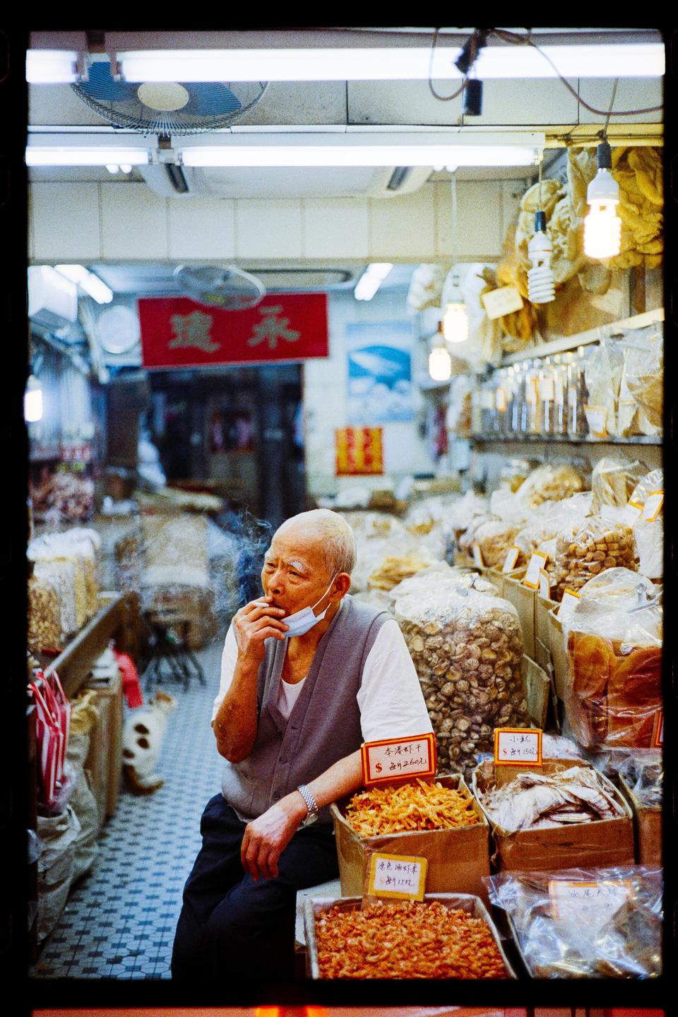 An old man with a face mask pulled down smokes a cigarette while sitting in a shop surrounded by bags and boxes of dried food.