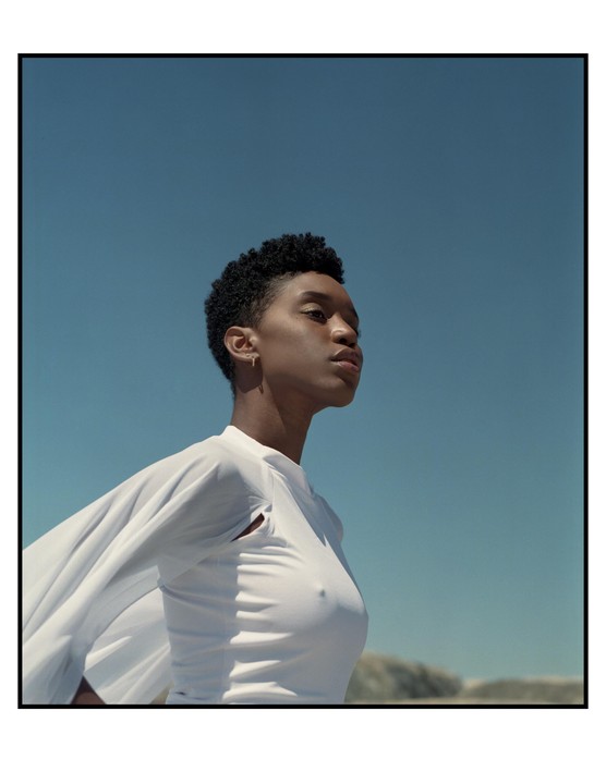 A woman with short curly hair wears a white top with flowing sleeves against a clear blue sky.
