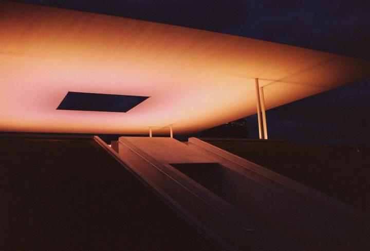 A large, illuminated ceiling with a rectangular opening is supported by columns above a ramp at night.