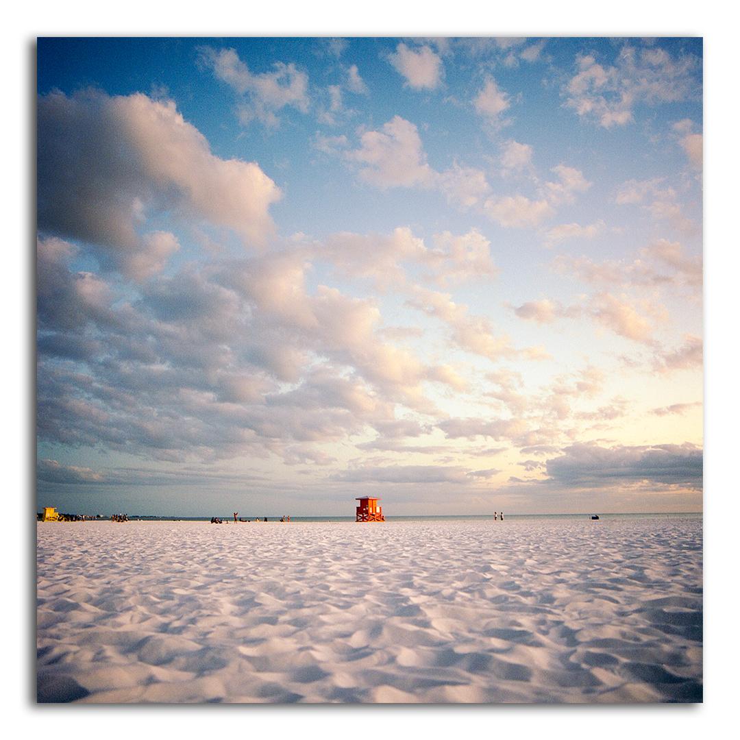 A wide expanse of white sand beach leads to the ocean under a cloudy sky at sunset.