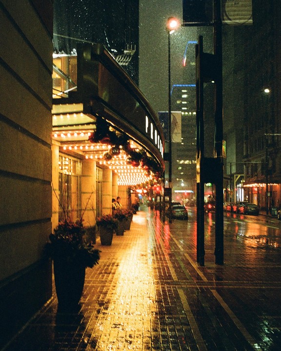 A street is illuminated by the lights of a building and streetlights at night while it snows.