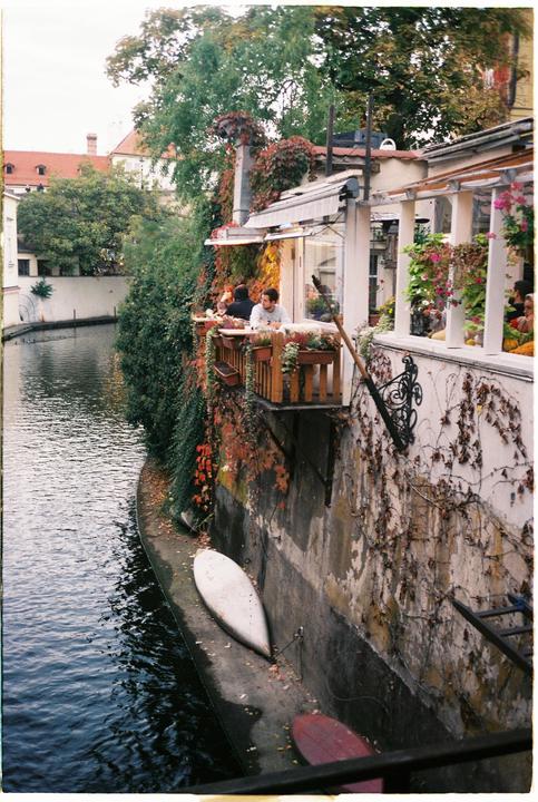 A restaurant with a balcony overlooking a canal is surrounded by lush green and red foliage.
