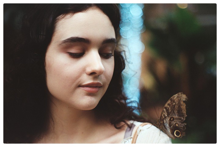 A butterfly rests on a woman's shoulder as she looks down with her eyes closed.