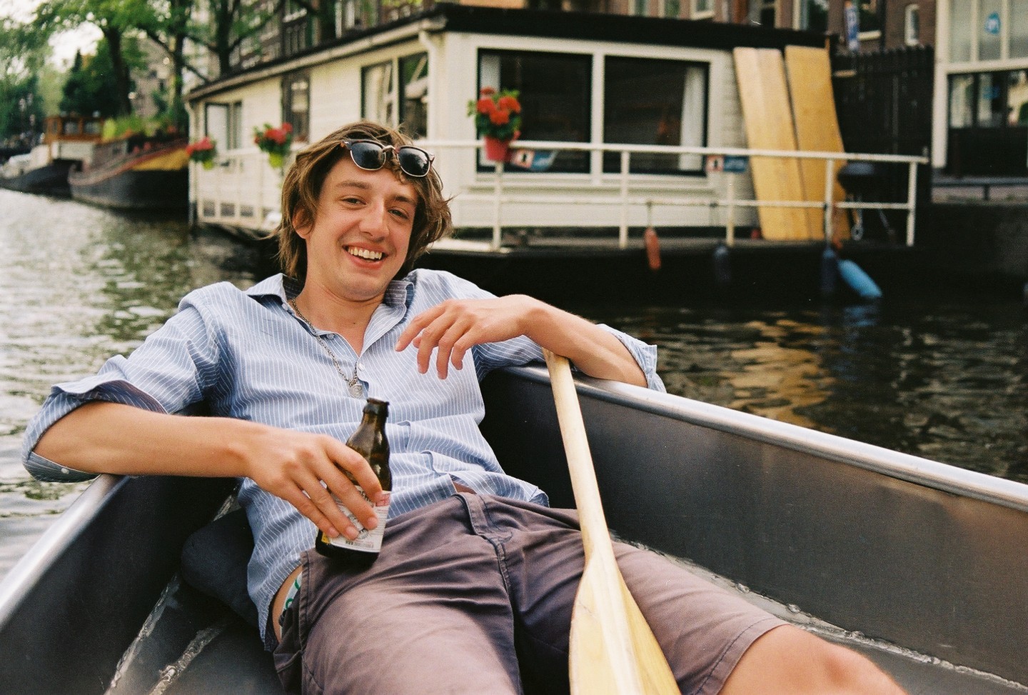 A man in sunglasses and a striped shirt sits in a canoe with a beer and a paddle, smiling at the camera with houseboats in the background.