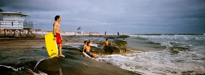 A man with a yellow boogie board stands on a rock at the beach while other children play in the water and a lifeguard tower stands in the background.