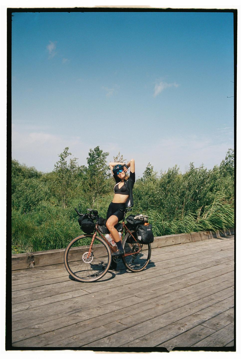 A woman in a helmet and sunglasses sits on a bicycle on a wooden boardwalk next to green foliage under a clear sky.