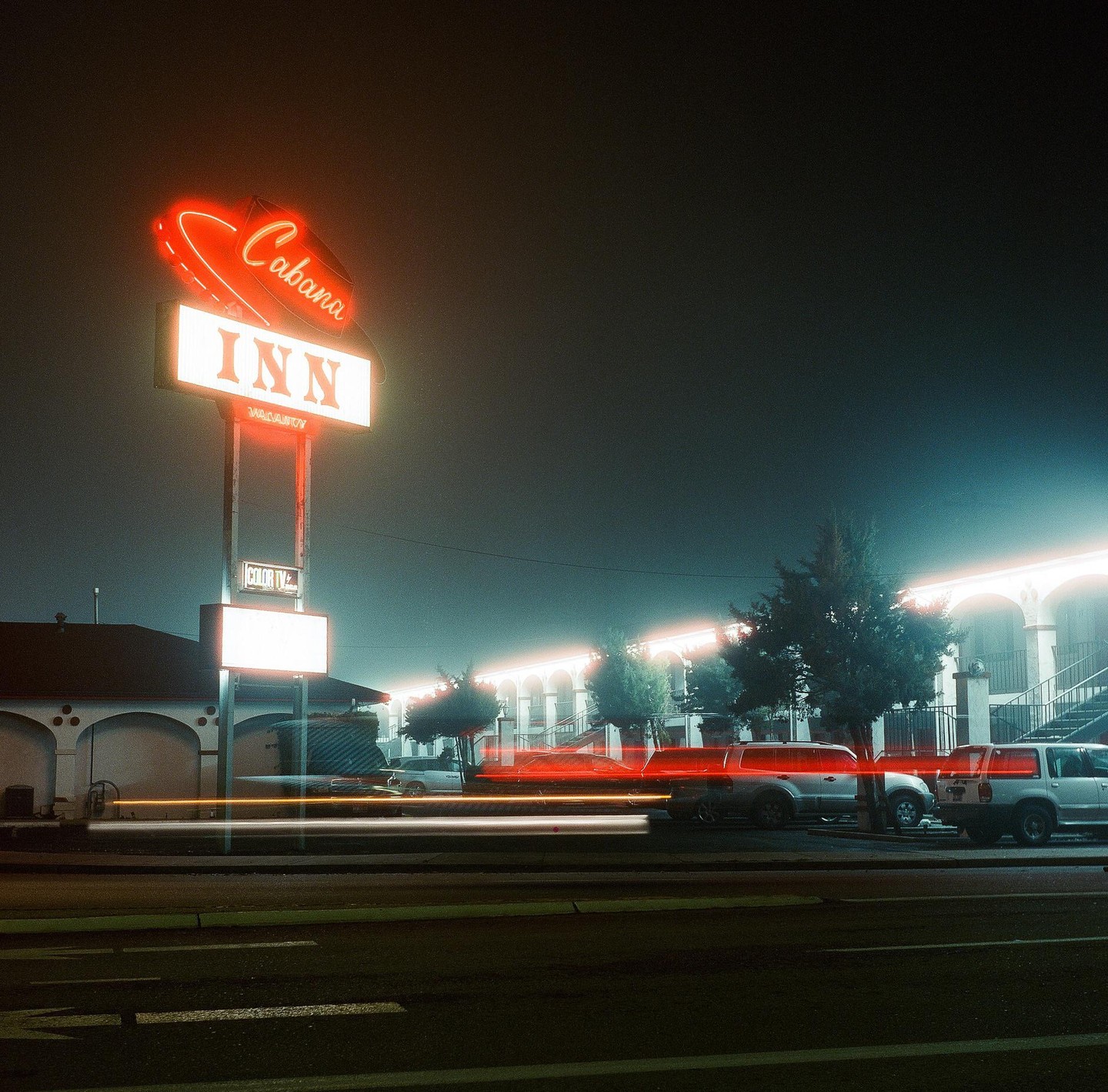 A neon sign for the Cabana Inn stands beside a street with light trails from passing vehicles at night.