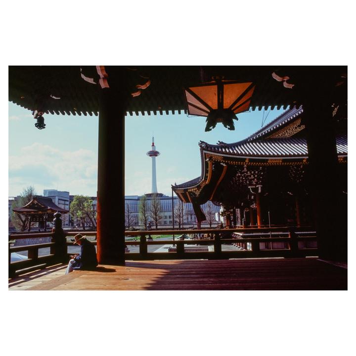 A man sits on a wooden deck of a temple with the Kyoto Tower visible in the distance.