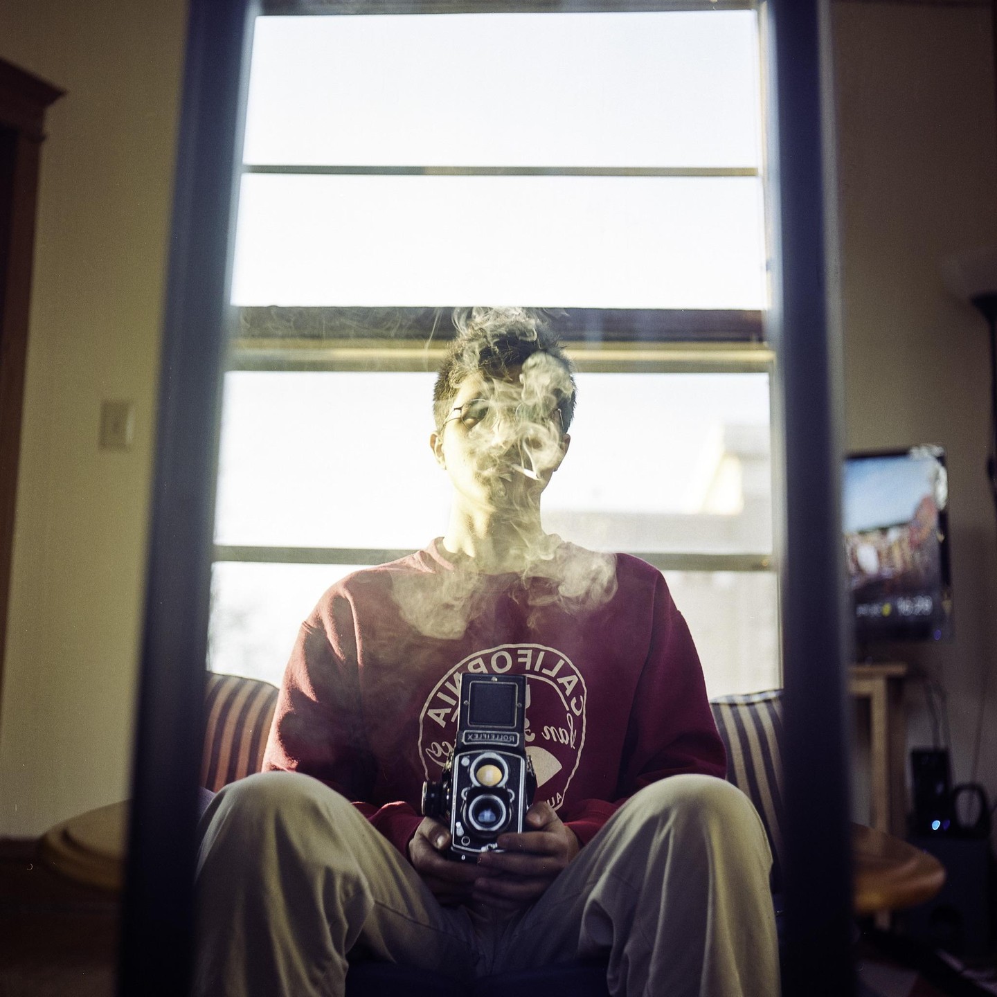 A man sits in a chair, holding a camera, with smoke around his head, reflected in a mirror.