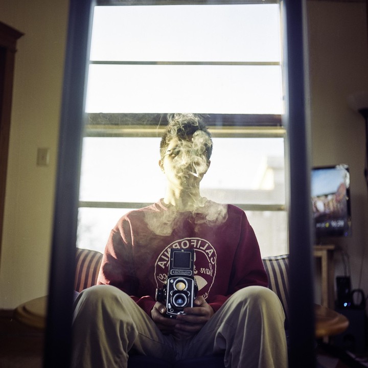 A man sits in a chair, holding a camera, with smoke around his head, reflected in a mirror.